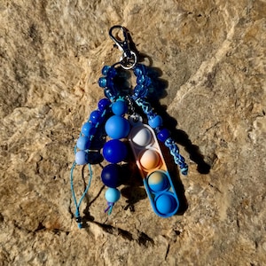 May include: A keychain featuring a silver-tone lobster clasp, a collection of blue and light blue spherical beads, and a rectangular silicone sensory toy with a gradient of white, orange, and blue circular push-pop bubbles.