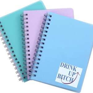 May include: Three spiral bound notebooks with colourful covers. The top notebook is light blue, the middle notebook is lavender, and the bottom notebook is light green. The light blue notebook has a sticker on the front that says "Drink Up Bitch" with an arrow pointing up.