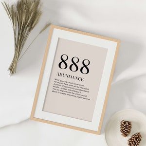 May include: Framed print with the number 888 and the word "ABUNDANCE" in a minimalist design. The print has a beige background and a white border, encased in a light wood frame. The text below describes abundance and opportunity.