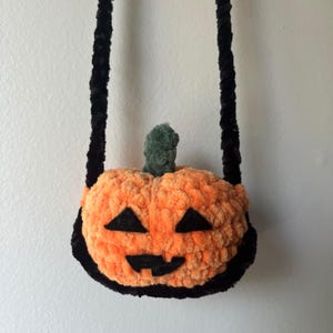 May include: A small orange crocheted pumpkin-shaped bag with a black strap. The pumpkin has a black face with eyes, a nose, and a mouth.