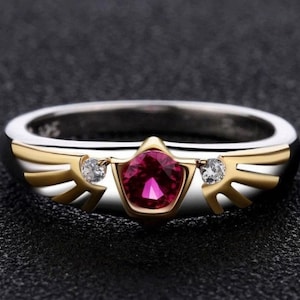 May include: A silver and gold ring with a central, faceted, deep red gemstone. The ring features gold wing-like accents on either side of the gemstone, and small, clear accent stones. The ring is set against a dark background.