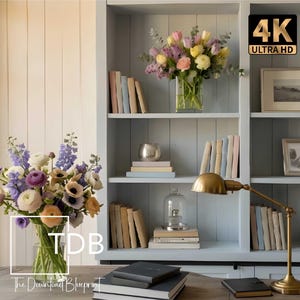 May include: A light blue wooden bookshelf with multiple shelves containing books, framed pictures, glass cloches, and two floral arrangements with purple, yellow, and white flowers. A brass desk lamp and black notebooks sit on the desk in front.