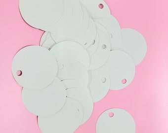 May include: A stack of white round paper tags with a hole punched in the center. The tags are on a pink background.