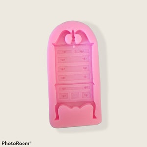 May include: Pink silicone mold for making a cake or dessert shaped like a tall, antique dresser with six drawers and four legs.
