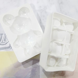 May include: Two white silicone molds, one shaped like a bear and the other with multiple rectangular cavities.