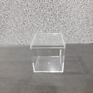 May include: A clear plastic cube-shaped box with a lid. The box is empty.