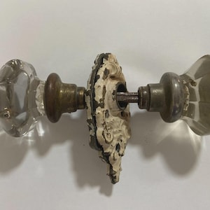 May include: Antique door hardware set featuring two faceted glass doorknobs with brass fittings. The set includes a decorative, off-white, ornate backplate. The hardware shows signs of age and wear, adding to its vintage appeal.