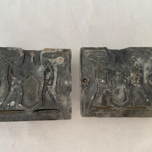 May include: Two antique metal molds, each with a rectangular shape and a detailed relief of a soldier. The molds are dark gray with signs of age and wear. The soldier figures are depicted with rifles and backpacks, suggesting a historical theme.