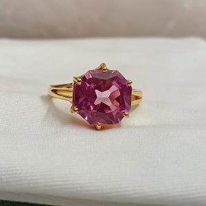 Puede incluir: Un anillo de oro con una gran gema rosa facetada. La gema octogonal está engastada en una banda de oro con un diseño de vástago dividido. El anillo se exhibe sobre una superficie blanca.