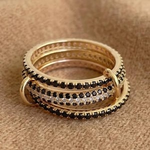 May include: A gold-toned ring set consisting of three interconnected bands. Two bands are encrusted with small black stones, while the central band features a row of clear white stones. The rings are linked together by a small gold-toned loop.