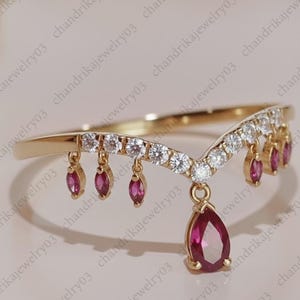 May include: A gold-toned ring with a V-shaped design. The ring features a row of small, clear gemstones and dangling ruby-colored teardrop-shaped gems. A larger ruby-colored teardrop gem hangs from the center of the V.