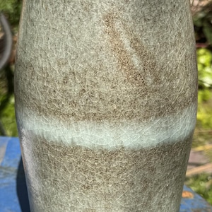 Könnte beinhalten: Eine hohe Keramikvase mit Craquelé-Glasur. Die Vase hat hauptsächlich eine gedämpfte grün-graue Farbe, mit einem helleren Band in der Mitte. Die Oberfläche hat eine leicht strukturierte Optik.