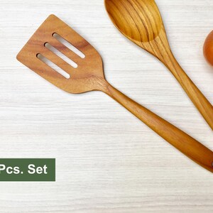 May include: A two-piece set consisting of a wooden slotted spatula and a wooden spoon, both featuring a natural light brown wood grain pattern. The set is displayed alongside one whole yellow onion and one whole red tomato. Text reads 2 Pcs. Set.