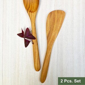 May include: A two-piece wooden kitchen utensil set consisting of one spoon and one flat-edged spatula, both featuring a natural light brown wood grain finish. A small purple clover leaf is positioned near the spoon handle. Text reads 2 Pcs. Set.