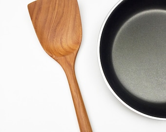 Peut inclure: Une spatule en bois avec une tête large et plate et un long manche fin doté d'un petit trou à l'extrémité, placée à côté d'une poêle à frire antiadhésive noire avec un bord argenté. Le grain du bois est visible sur toute la spatule.