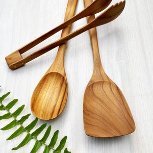 May include: A set of three wooden kitchen utensils including a pair of tongs, a rounded spoon, and a flat-edged spatula. Each piece features a natural light brown wood grain finish. The text "Set of 3 pcs." is printed in the bottom right corner.
