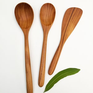 May include: A set of three wooden kitchen utensils consisting of two spoons and one flat spatula, all featuring a natural brown wood grain pattern. A single green leaf is positioned near the base of the utensils, and the text Set of 3 pcs. is visible.