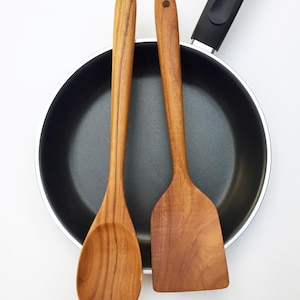 May include: A black non-stick frying pan with a long handle, paired with two light brown wooden kitchen utensils, a spoon and a flat spatula. The text "Set of 2 Pcs." is printed in the bottom right corner of the image, indicating the utensil count.
