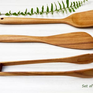 May include: A set of three light brown wooden kitchen utensils including a slotted spatula with a hole in the handle, a flat paddle spatula, and a pair of salad tongs with serrated edges. The text "Set of 3 pcs." is printed in the bottom right corner.