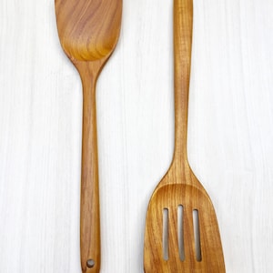 Könnte beinhalten: Zwei Küchenutensilien aus Holz, ein Spatel und ein Schöpflöffel mit Schlitzen, beide mit einer natürlichen Holzoptik. Der Spatel hat einen abgerundeten Kopf und der Schöpflöffel mit Schlitzen hat einen Schlitz-Kopf mit drei Schlitzen.