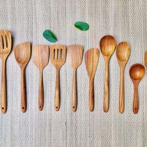 May include: A collection of twelve light brown wooden kitchen utensils arranged in a row, including various spatulas, slotted turners, spoons, a ladle, and a pair of tongs, all featuring smooth, polished surfaces and small holes at the handle ends.