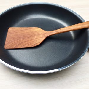 Könnte beinhalten: Eine schwarze Bratpfanne mit einem Holzspatel darin. Der Spatel ist aus hellbraunem Holz gefertigt. Die Pfanne hat einen silbernen Rand und einen schwarzen Griff. Der Pfannendurchmesser beträgt ca. 25 cm.