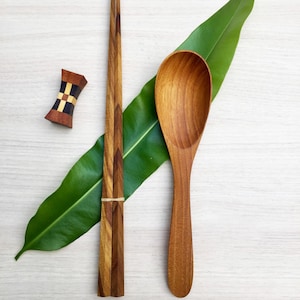 May include: A wooden spoon and a pair of wooden chopsticks with a small, rectangular, multi-colored wooden chopstick rest featuring a geometric pattern. The set is arranged on a green leaf, and the text Chopsticks & Spoon Set is visible.