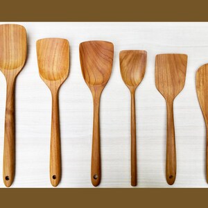 May include: A set of six wooden spatulas arranged in a row, each featuring a distinct grain pattern and a light brown finish. Every spatula has a flat, wide head and a long handle with a small circular hole at the base for hanging purposes.