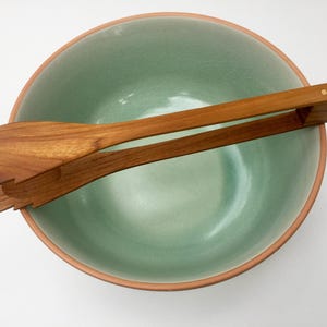 May include: A round ceramic bowl with a light mint green interior and a terracotta-colored rim, paired with a set of wooden salad tongs featuring a natural grain finish and a hinged design resting across the top of the bowl.
