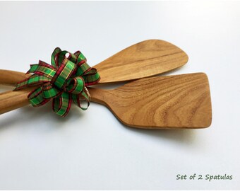 May include: A set of two light brown wooden spatulas with visible wood grain, tied together with a decorative green and red plaid ribbon bow. The text Set of 2 Spatulas is printed in the bottom right corner of the image.