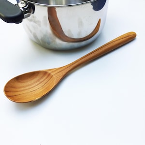 Könnte beinhalten: Ein Holzlöffel mit abgerundeter Schüssel und langem Griff, der auf einer weißen Oberfläche neben einem Edelstahltopf liegt. Der Löffel ist hellbraun mit sichtbarer Holzmaserung. Der Topf hat einen schwarzen Griff.