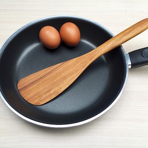 May include: A black non-stick frying pan with a silver rim and a black handle, containing two brown chicken eggs and a wooden spatula with a flat, wide blade and a long, tapered handle resting inside the pan.