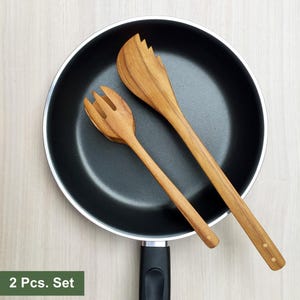 May include: A black non-stick frying pan containing a two-piece wooden salad serving set. The set includes a fork and a wide, serrated-edge server, both featuring a natural wood grain finish. The text 2 Pcs. Set is visible in the corner.