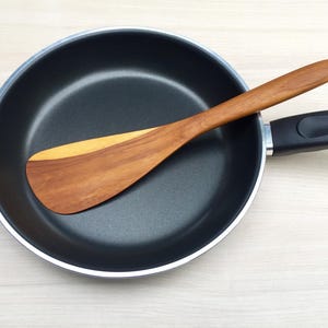 May include: A round black non-stick frying pan with a silver rim and a black handle. A flat wooden spatula with a two-toned brown grain pattern is resting diagonally across the interior surface of the pan, extending past the edge.