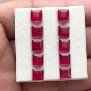 May include: Ten square-cut, vibrant red gemstones are arranged in two parallel rows on a white surface. The gemstones have a polished, reflective surface, and are likely rubies. The gemstones are set against a white background.