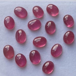 May include: A collection of oval-shaped, polished gemstones in varying shades of pink and red. The gems are smooth and reflective, with a glossy surface. They are arranged on a white surface, showcasing their color and clarity.