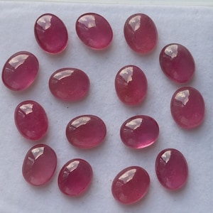May include: A collection of oval-shaped, polished ruby gemstones. The gems are a deep, translucent red with a glossy finish, reflecting light. The stones are arranged on a white surface, showcasing their color and clarity.