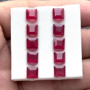 Peut inclure: Dix pierres précieuses carrées, rouge foncé, disposées en deux colonnes de cinq sur une surface blanche. Les pierres précieuses ont une surface polie et réfléchissante, et sont probablement des rubis. Les gemmes sont présentées sur un fond neutre.