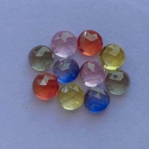 Puede incluir: Una colección de piedras preciosas facetadas en varios colores, incluyendo rosa, naranja, amarillo, verde y azul. Las piedras preciosas son redondas y parecen pulidas, con una superficie reflectante. Las gemas están dispuestas sobre un fondo morado claro.
