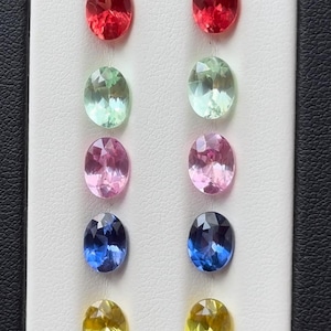 Puede incluir: Una variedad de gemas de corte ovalado en varios colores, incluyendo rojo, verde, rosa, azul y amarillo. Las gemas se exhiben sobre una superficie blanca con un borde negro, mostrando sus facetas.