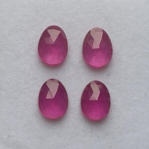 May include: Four oval-shaped, faceted gemstones in a vibrant magenta hue. The gems are arranged in a square pattern against a neutral background. Each stone has a polished, reflective surface, showcasing the cut's precision.