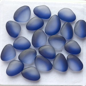 May include: A collection of smooth, blue sea glass pieces. The pieces vary in shape, with rounded and triangular forms. The color transitions from a deep blue to a lighter, frosted blue, creating a gradient effect. The sea glass is arranged on a white surface.