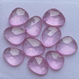May include: A collection of ten faceted, light pink gemstones. The gemstones are in various shapes, including teardrop and rounded triangle. Each stone has multiple facets that catch the light, creating a sparkling effect. The stones are arranged closely together.