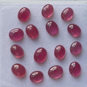 May include: A collection of oval-shaped, polished ruby gemstones. The gems are a deep, rich red color with a glossy surface. They are arranged on a white surface, showcasing their vibrant hue and clarity. The gemstones are ready for jewelry making.