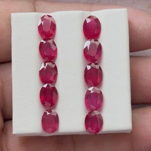Puede incluir: Una colección de diez rubíes de talla ovalada, cada uno mostrando un tono rojo intenso. Las piedras preciosas están dispuestas en dos filas verticales de cinco, sobre un fondo blanco. Los rubíes están facetados, reflejando la luz.