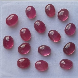 May include: A collection of oval-shaped, polished ruby gemstones. The gems are a deep, rich red color with a smooth, reflective surface. The gemstones are arranged on a white surface, showcasing their natural beauty and potential for jewelry making.