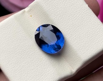 Puede incluir: Una gema facetada de forma ovalada con un tono azul profundo. La gema se encuentra sobre una superficie blanca, mostrando su claridad y brillo. El corte de la piedra crea múltiples superficies reflectantes.