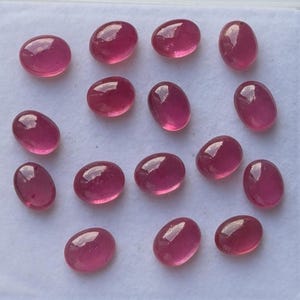 May include: A collection of oval-shaped, polished ruby gemstones. The gems are a deep, translucent red color, with a smooth, reflective surface. The stones are arranged on a white surface, showcasing their vibrant color and clarity.