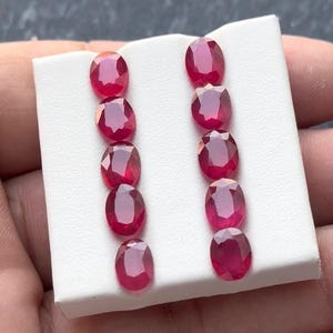 May include: Ten oval-cut, deep red rubies are arranged in two columns on a white surface. The gemstones have a polished, reflective surface, showcasing their rich color and clarity. The rubies are likely for jewelry making.