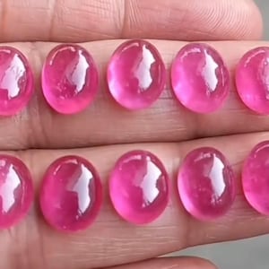 May include: Ten oval-shaped, polished pink gemstones arranged in two rows. Each stone features a smooth, rounded cabochon cut with a reflective surface, displaying a vibrant, consistent magenta-pink hue across all ten individual pieces.